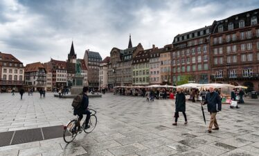 Place centrale de Strasbourg avec des bâtiments historiques, une statue équestre, des stands de marché et des passants. Un cycliste au premier plan sur une place pavée sous un ciel nuageux.