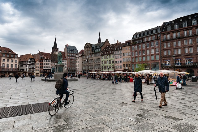 Place centrale de Strasbourg avec des bâtiments historiques, une statue équestre, des stands de marché et des passants. Un cycliste au premier plan sur une place pavée sous un ciel nuageux.