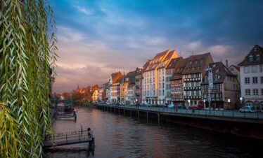 Canal de Strasbourg au crépuscule. Maisons anciennes alignées le long de l'eau, fleurs colorées au premier plan. Lumière dorée sur les façades et reflets dans le canal calme.