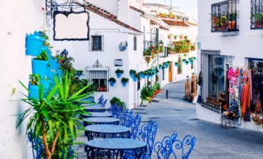 Vue sur une ruelle entre les maisons d'une blancheur immaculée. Les tables rondes en fer forgé et les chaises invitent à s'asseoir