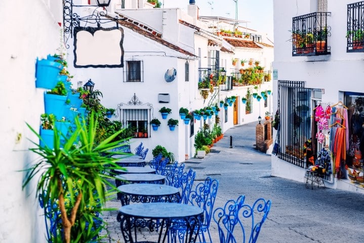 Vue sur une ruelle entre les maisons d'une blancheur immaculée. Les tables rondes en fer forgé et les chaises invitent à s'asseoir